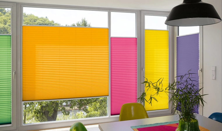 Verschiedenfarbene Plissees beschatten aneinanderliegende Fenster verschiedener Größen.