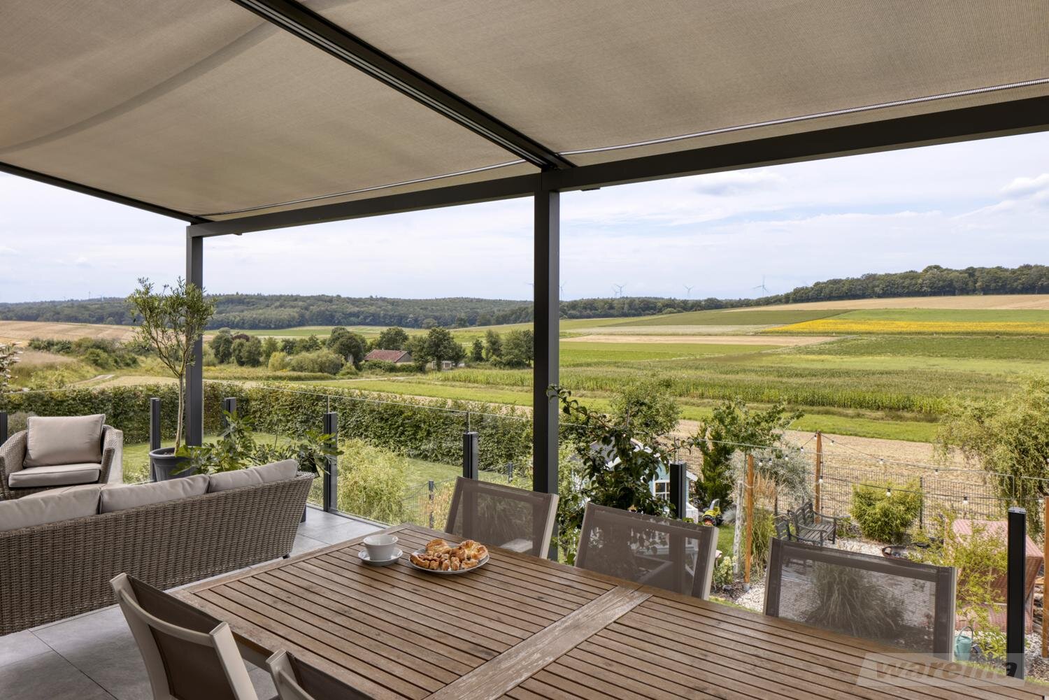 Blick über große Wiesenflächen von einer Terrasse mit Sitzgelegenheiten geschützt von einer Markise.