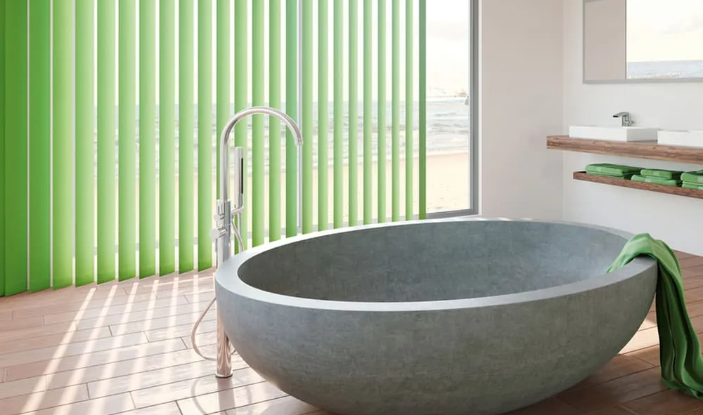 Eine halbrunde Badewanne aus Beton in einem Badezimmer. Grüne Handtücher setzen farbliche Akzente und die grüne Vertikaljalousie passt im Farbton.
